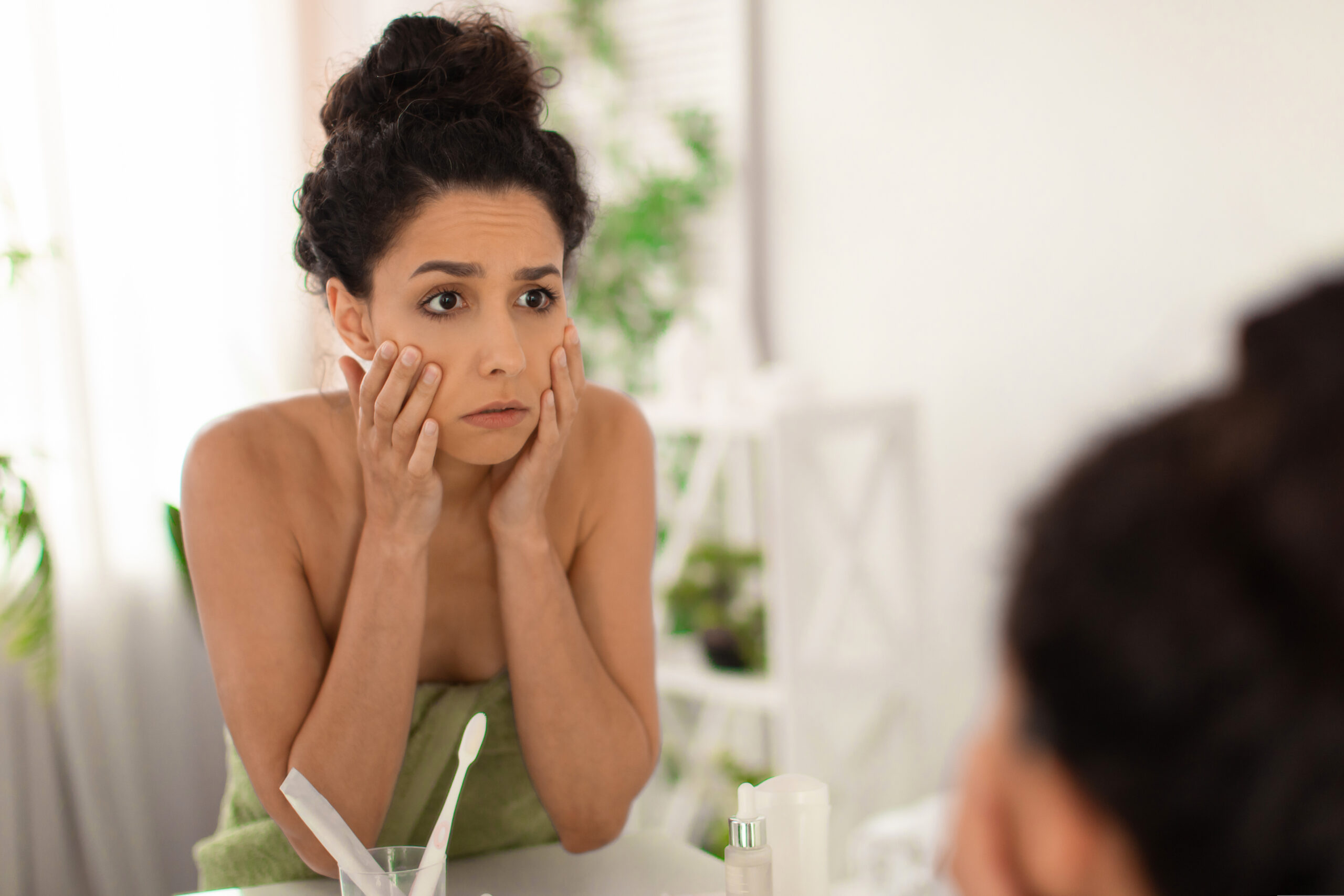 Upset young woman checking first wrinkles around her eyes, feeling tired or stressed, looking in mirror